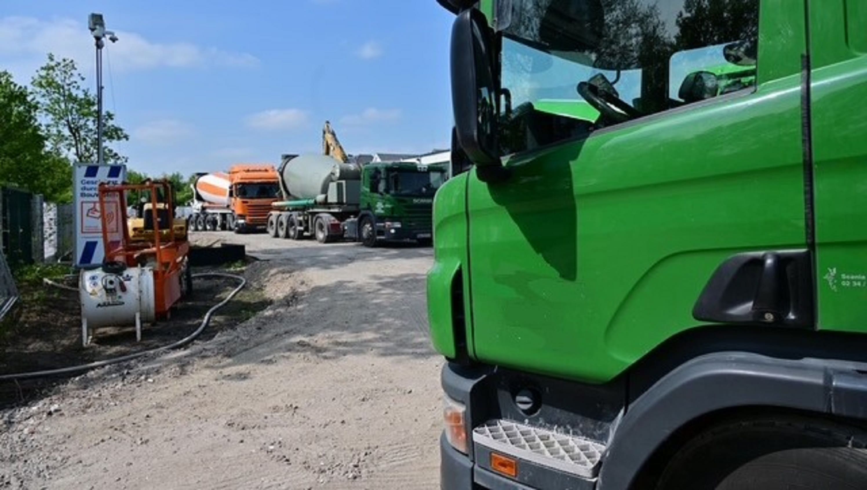 Mehrere Betonmischer auf Baustelle, vorne grüner LKW, daneben Bautechnik und Hinweisschild zu Sicherheitsregeln.